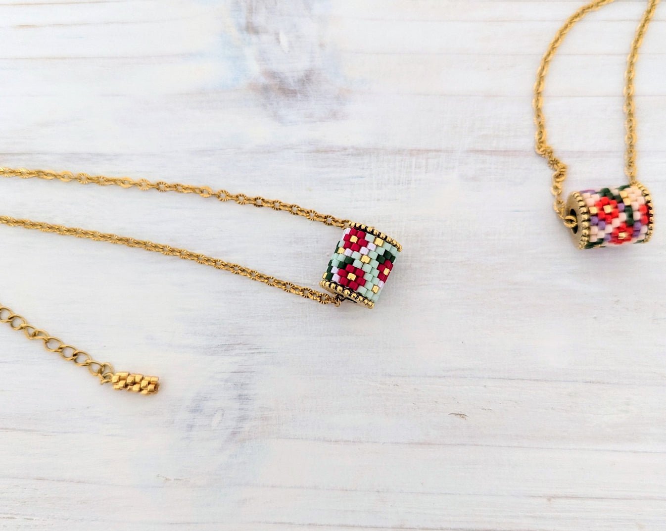 Gold chain necklace with a small cylindrical bead pendant featuring a colorful geometric pattern of red, green, black, and white beads, displayed on a light wood background.
