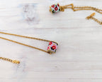Gold chain necklace with a small square bead pendant featuring a colorful floral pattern in red, pink, white, and green, displayed on a light wood background.