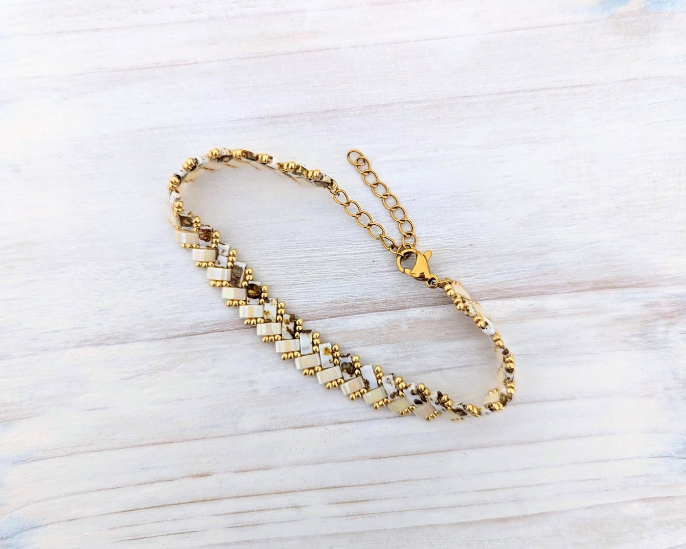 A gold-toned beaded chain bracelet with alternating white and gold square beads, displayed on a light-colored wooden surface.