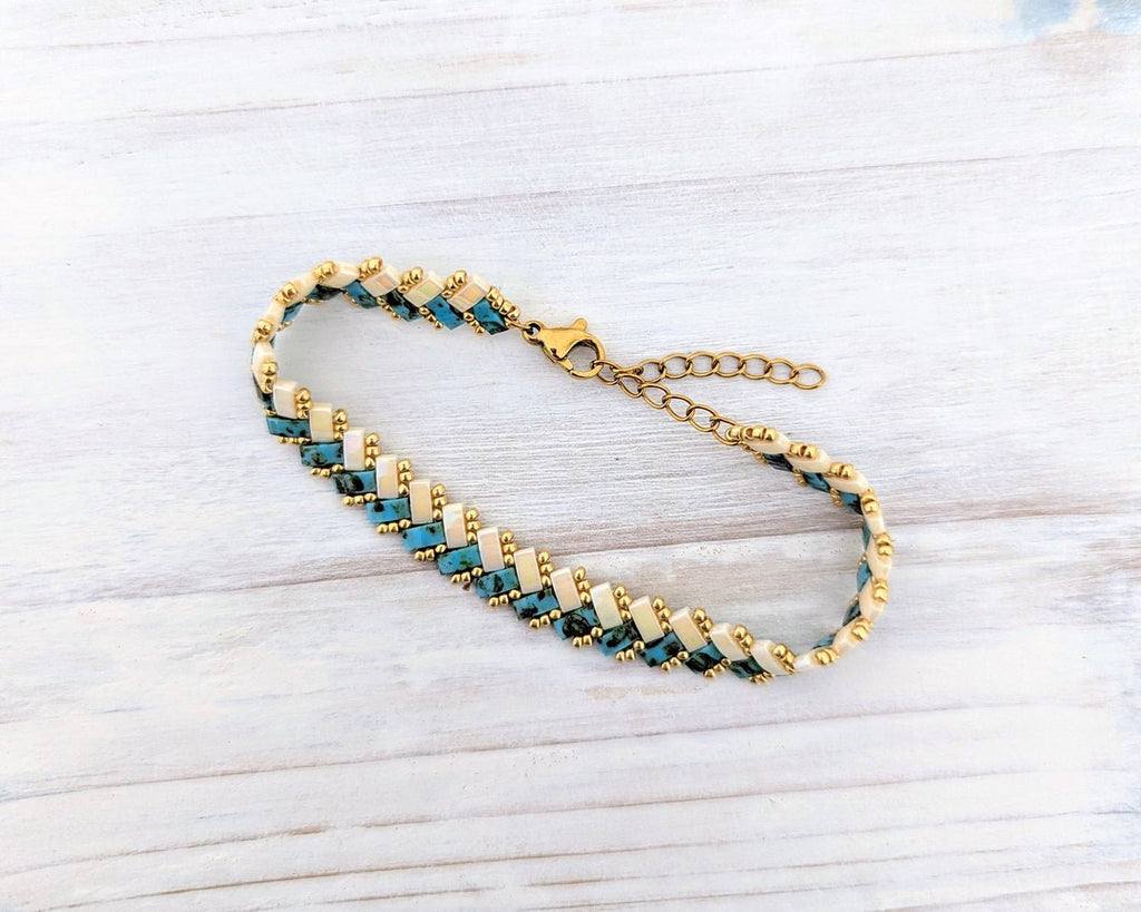 A gold chain bracelet with alternating rows of small teal blue and gold beads, displayed on a light gray wood background.