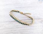 A gold chain bracelet with alternating rows of small teal blue and gold beads, displayed on a light gray wood background.