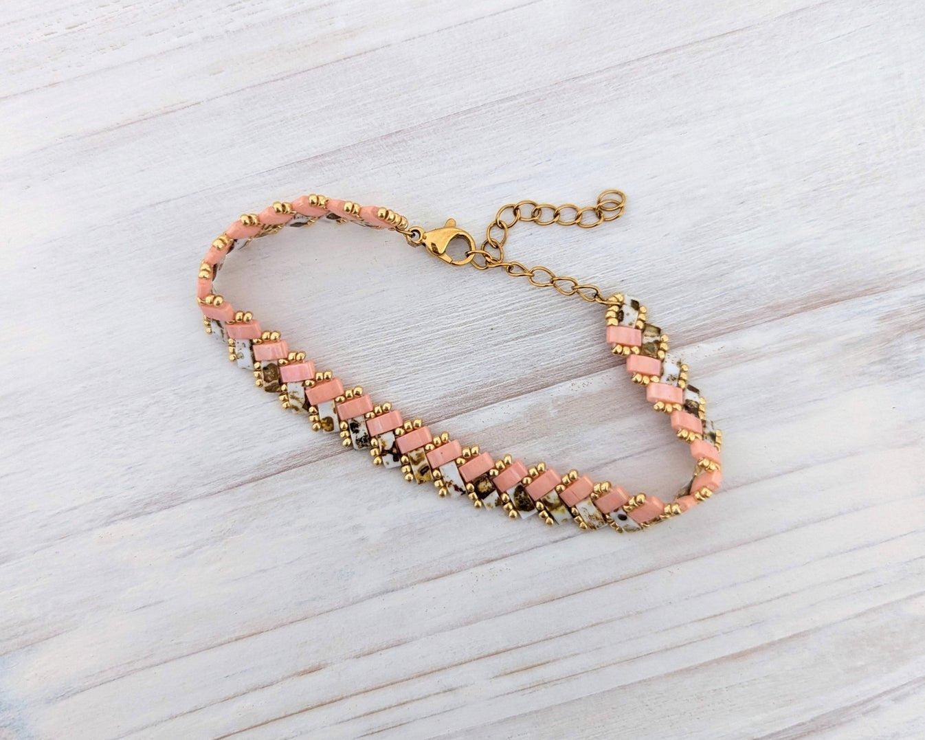 A handmade beaded bracelet with pink and white beads, gold spacer beads, and a gold clasp, displayed on a light gray wood background.