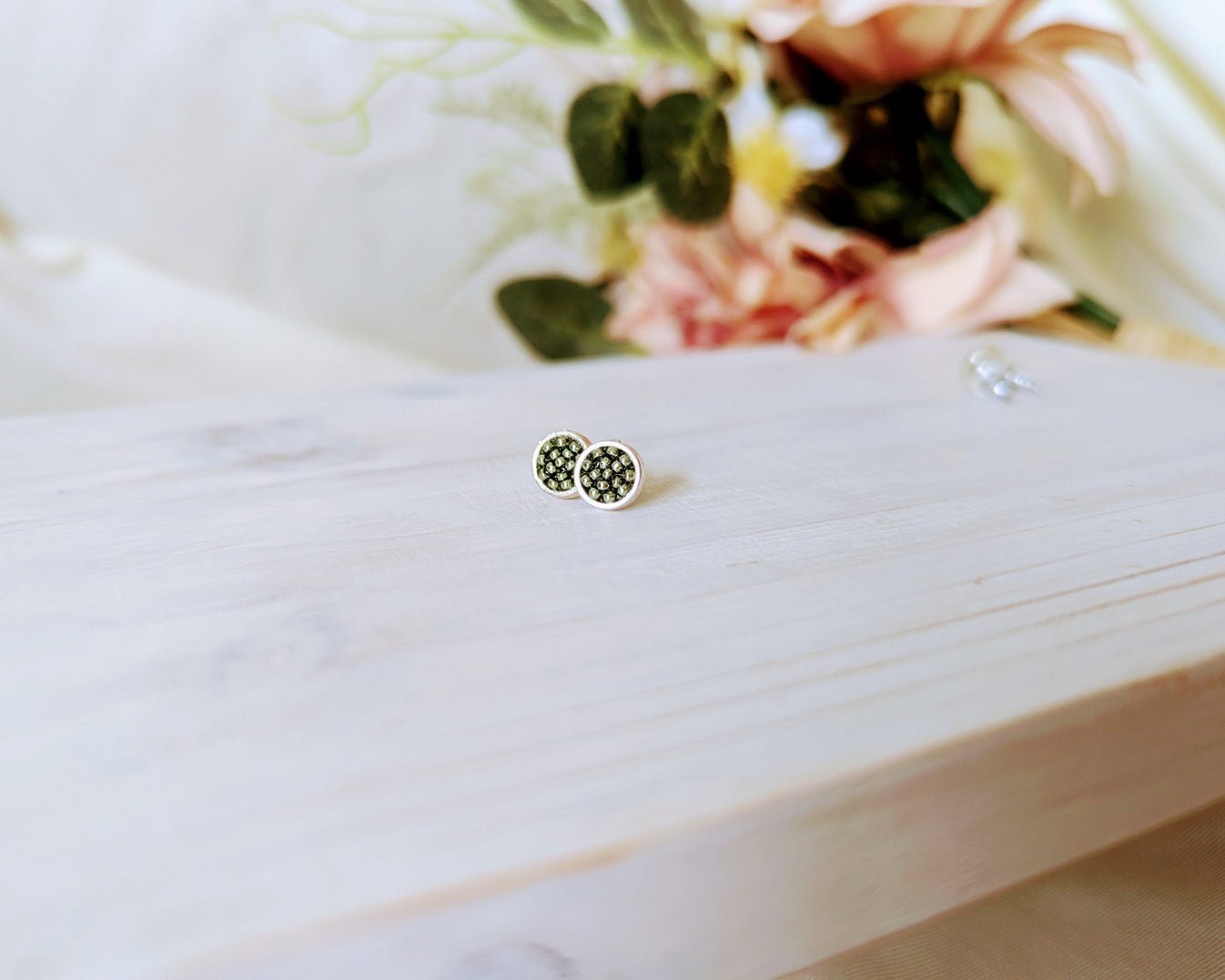 A pair of round stud earrings with a black and white textured pattern and silver borders, resting on a light-colored wooden surface with a soft-focus floral background.