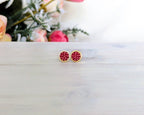 A pair of gold stud earrings with round red beaded centers, resting on a light gray wooden surface with a blurred floral background.