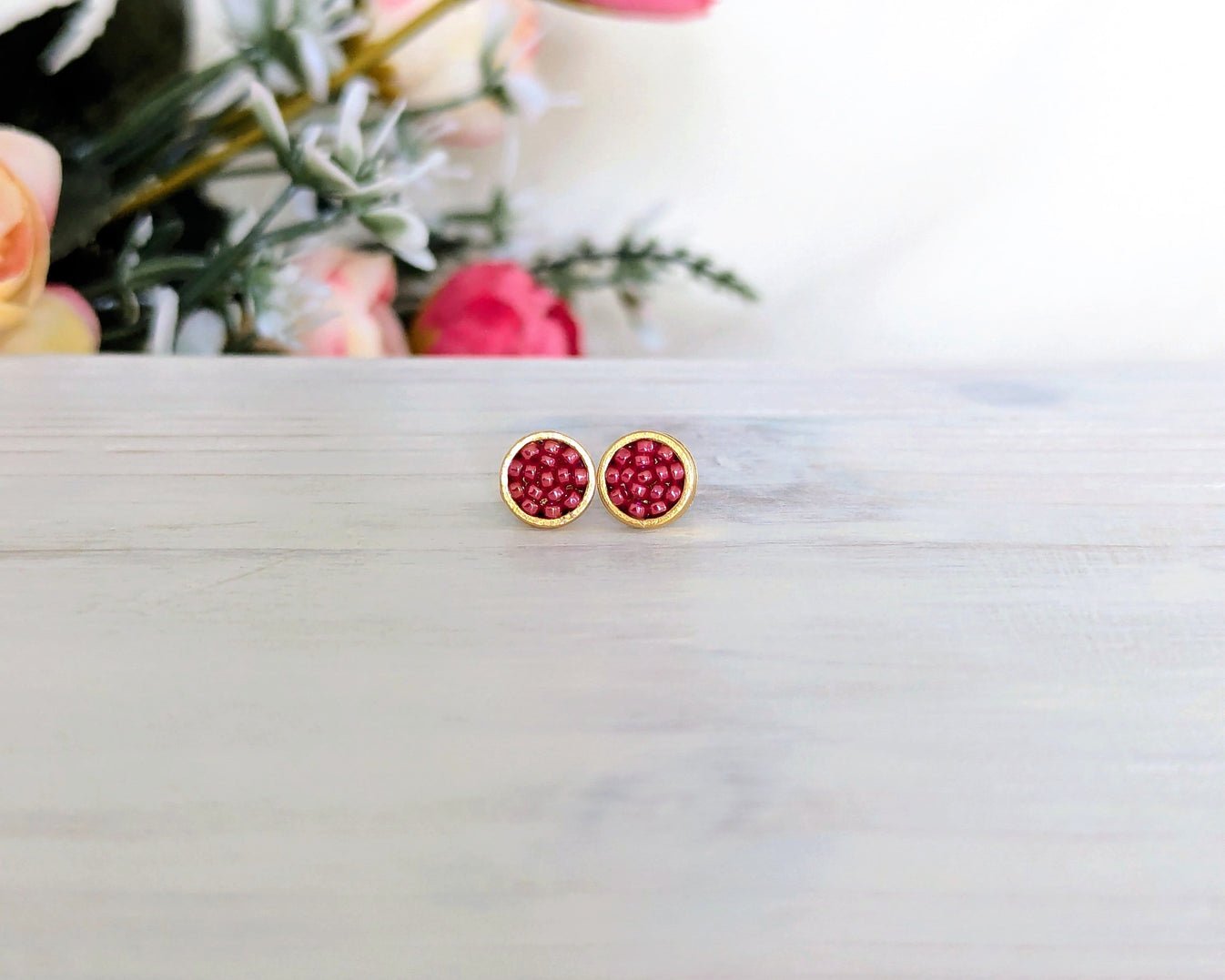 A pair of gold stud earrings with round red beaded centers, resting on a light gray wooden surface with a blurred floral background.
