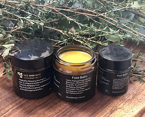 Three skincare jars arranged on a wooden surface with green leaves behind them: a black jar labeled Face Wash, an open black jar showing yellow balm labeled Face Balm, and a black jar labeled Body Wash.