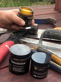 A hand holds a small amber jar of hair product above two black jars labeled 'HONEY THICK HAIR' on a workbench with tools, including a hammer and a grater.