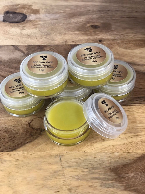 Several small clear plastic jars of beeswax lip balm with gold labels are arranged on a rustic wooden surface. One jar in the foreground is open, showing the yellow-green balm inside.