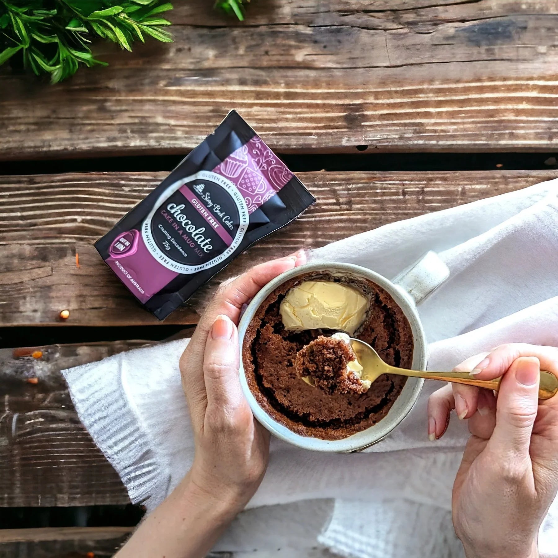 Chocolate mug cake with a scoop taken out, held by hands over a wooden surface with a package of chocolate mix.