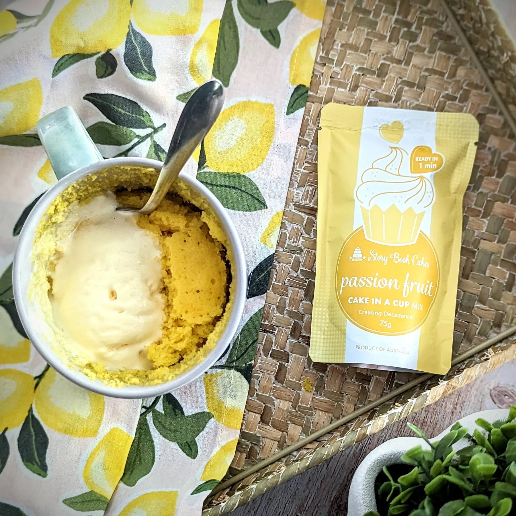A yellow and white pouch of Sunny Day Cakes passion fruit cake mix sits next to a white mug containing a cooked cake with a scoop of vanilla ice cream, styled on a lemon-print cloth and woven tray.