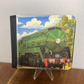 A bi-fold wallet featuring a colorful illustration of a green steam locomotive train under a blue sky with white clouds, displayed on a clear stand on a wooden surface against a white wall.