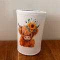 A white neoprene cup sleeve featuring a cute illustration of a fluffy orange-brown Highland cow with a sunflower crown, sitting on a wooden table against a plain wall.