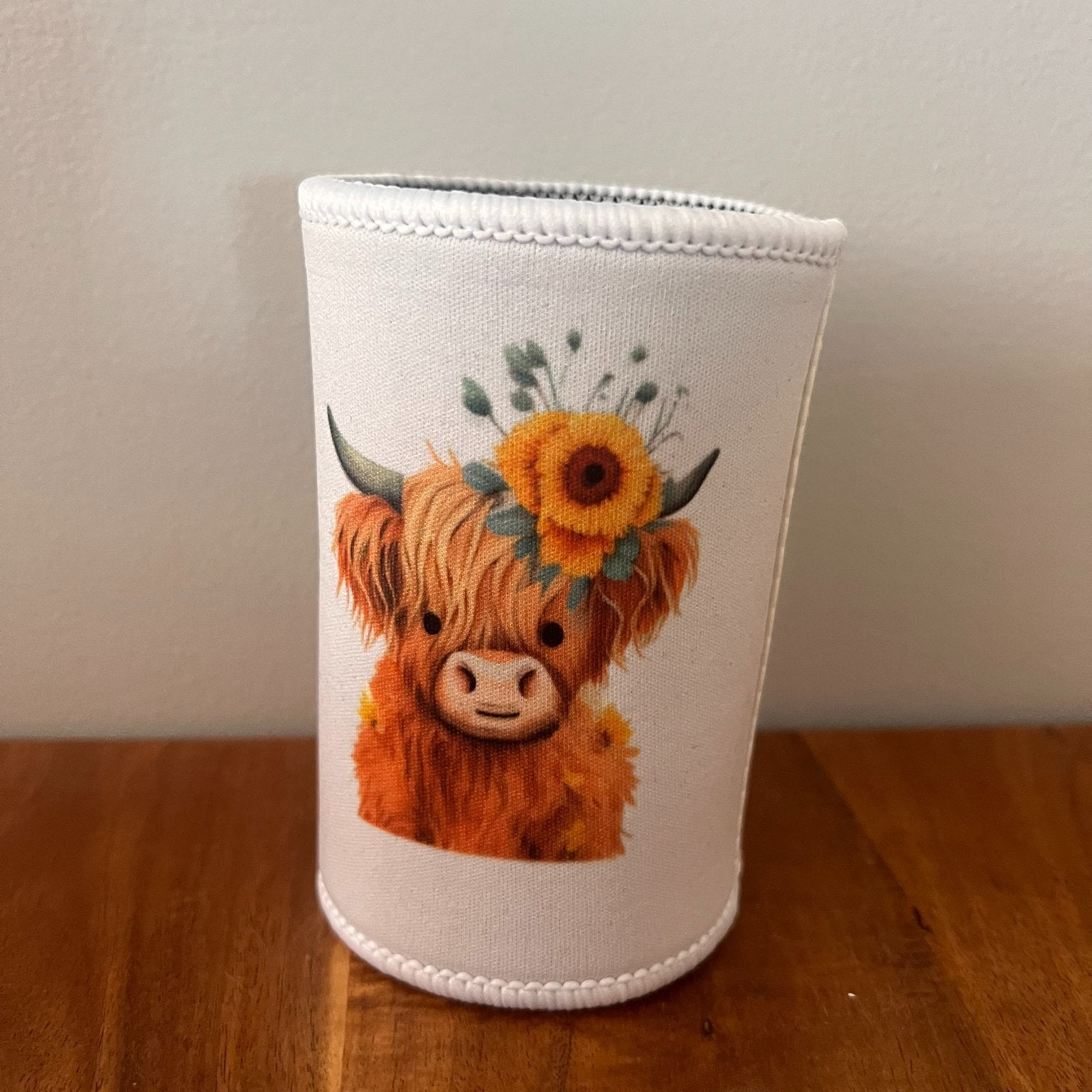 A white neoprene cup sleeve featuring a cute illustration of a fluffy orange-brown Highland cow with a sunflower crown, sitting on a wooden table against a plain wall.