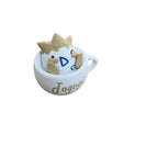 A small white ceramic cup with the word 'Jogeni' printed on the side, holding a beige clay-like character with a crown and blue geometric face details, placed on a wooden tabletop.