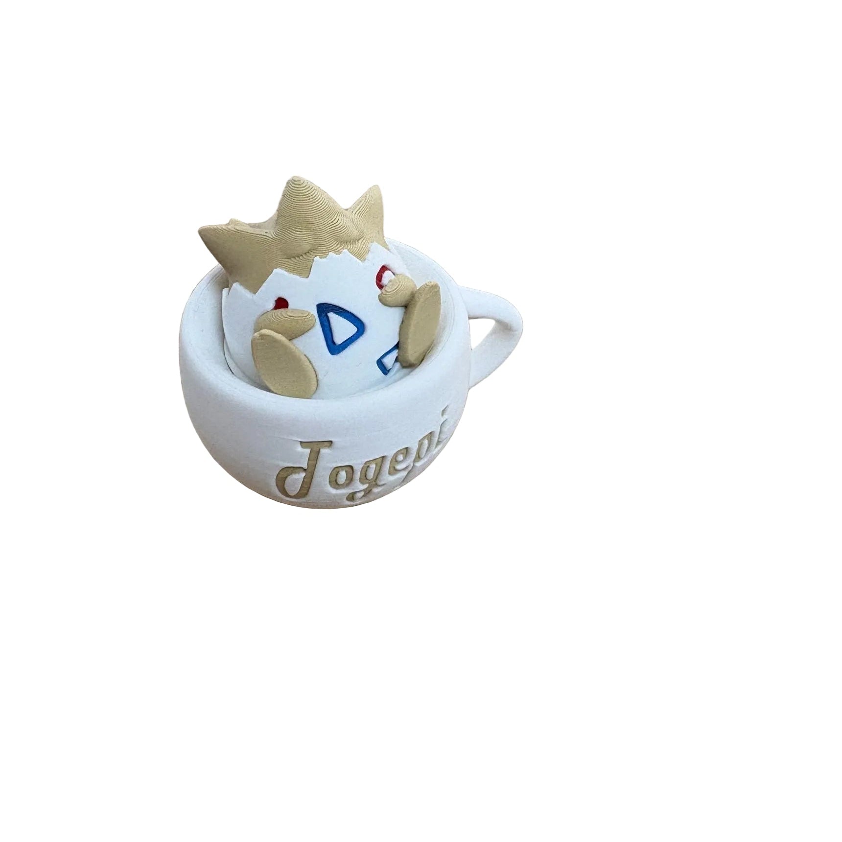 A small white ceramic cup with the word 'Jogeni' printed on the side, holding a beige clay-like character with a crown and blue geometric face details, placed on a wooden tabletop.